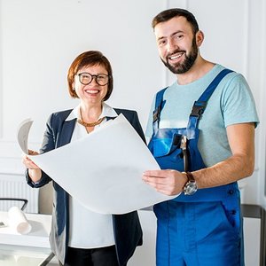 EIne Frau und ein Mann der Arbeitskleidung trägt stehen in einem Raum und halten eine Zeichnung fest